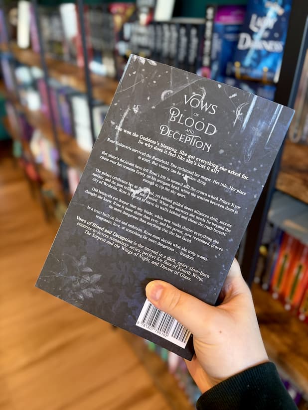 A hand holding the back of a copy of 'Vows of Blood and Deception' by Aria Ashbrook with a dark, fantasy-themed design and icicles and frost creep in from the edges of the cover.