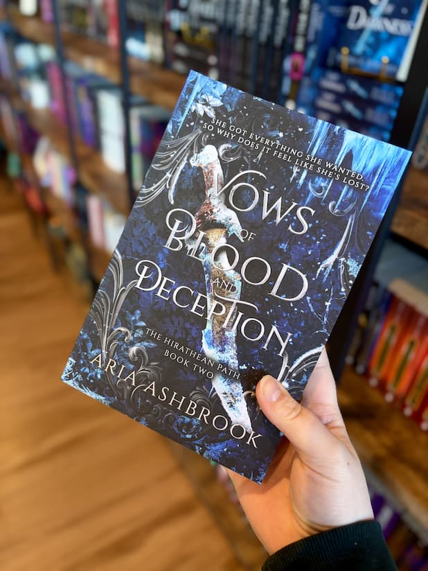 A hand holding a copy of 'Vows of Blood and Deception' by Aria Ashbrook with a dark, fantasy-themed design. A knife sits frozen in ice in the middle and icicles and frost creep in from the edges of the cover.