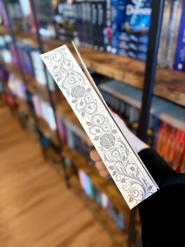 A hand holding a copy of 'Vows of Blood and Deception' by Aria Ashbrook on the side, showing sprayed edges of flowers and leaves.