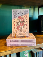 A copy of the book 'Want It All' by Bollie Hartwright stands upright on top of two stacked copies of the same book, displayed on a wooden shelf against a teal background.