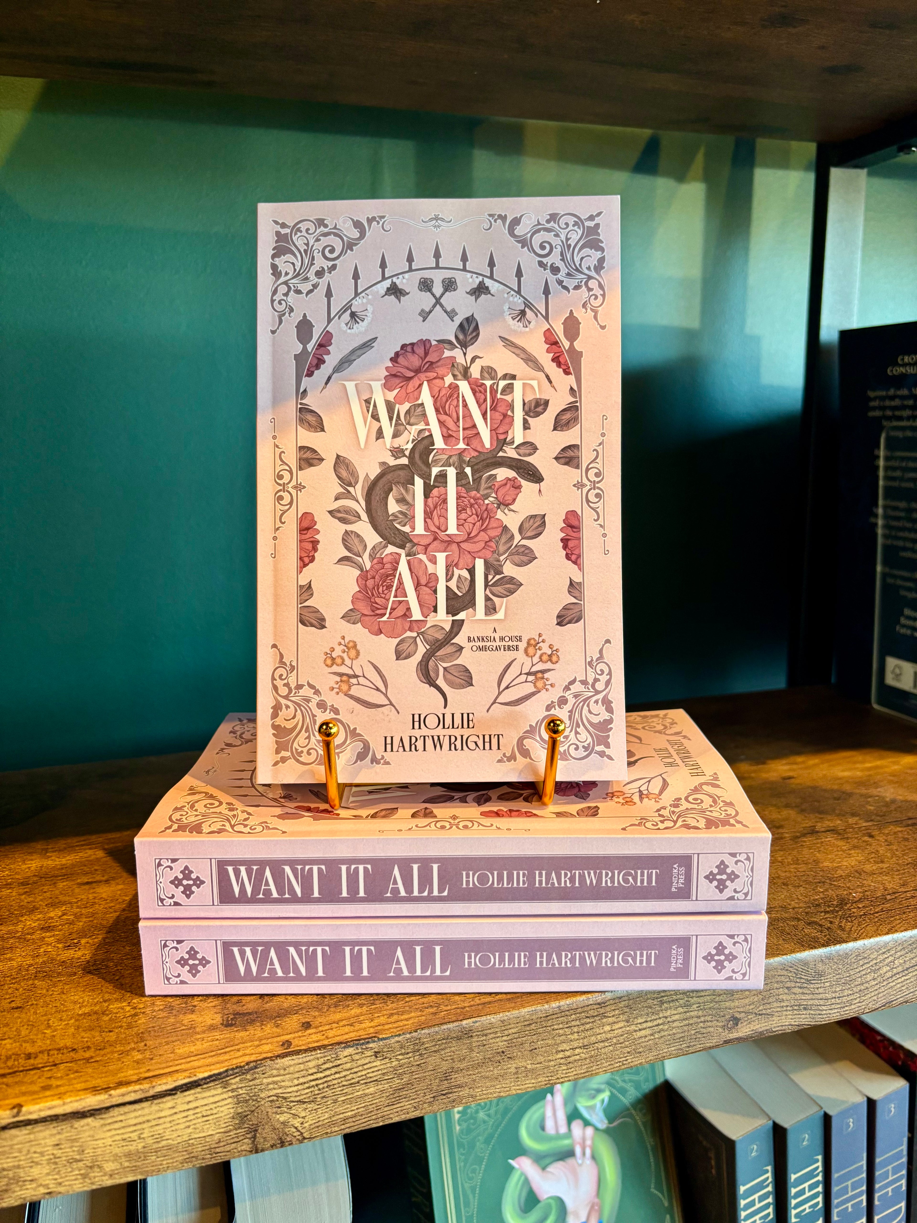 A copy of the book 'Want It All' by Bollie Hartwright stands upright on top of two stacked copies of the same book, displayed on a wooden shelf against a teal background.