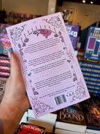 A hand holds a light pink book cover of Want It All by Hollie Hartwright with ornate dusty pink floral illustrations and text, photographed in a bookstore with other books blurred in the background.