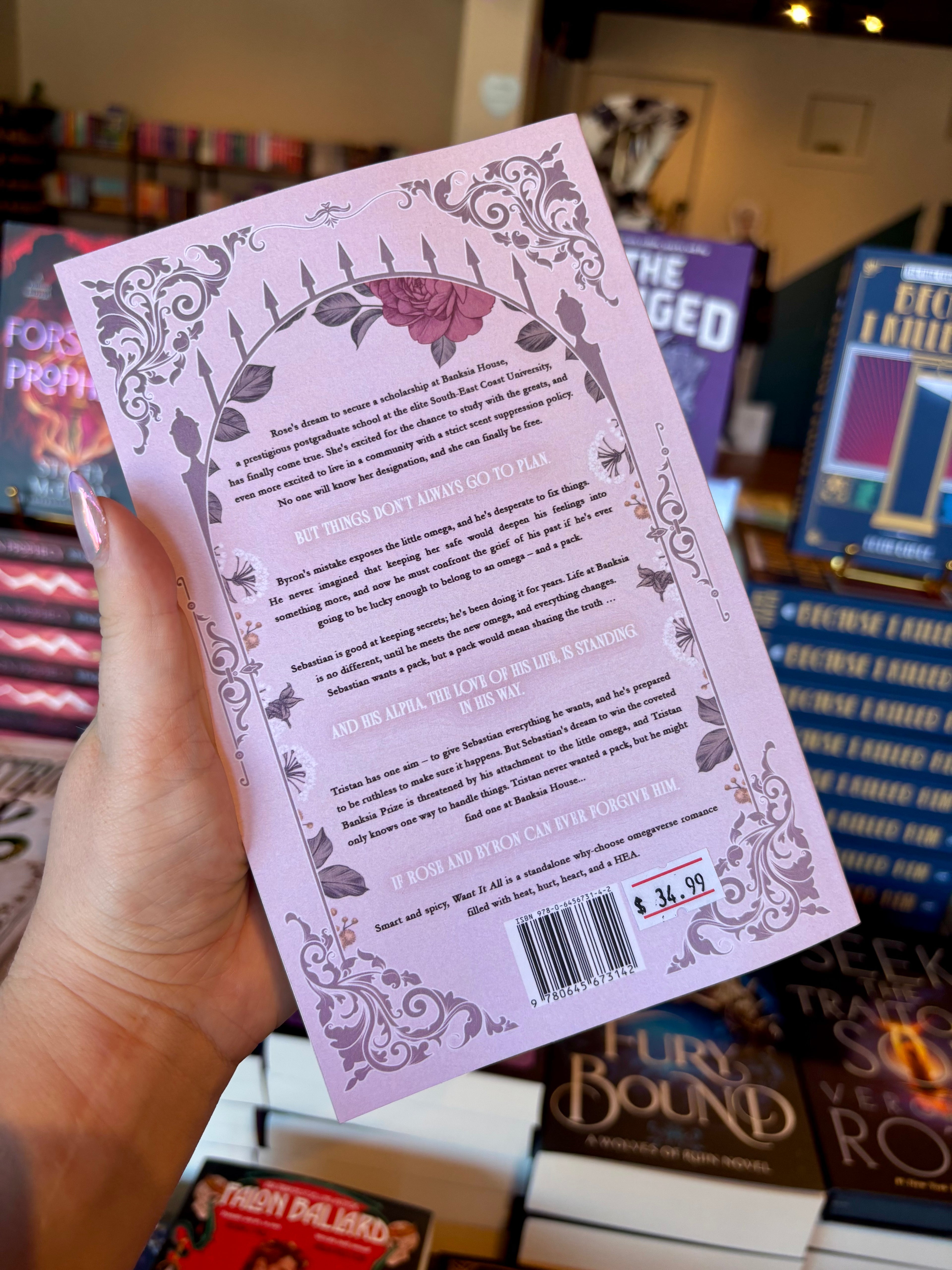 A hand holds a light pink book cover of Want It All by Hollie Hartwright with ornate dusty pink floral illustrations and text, photographed in a bookstore with other books blurred in the background.