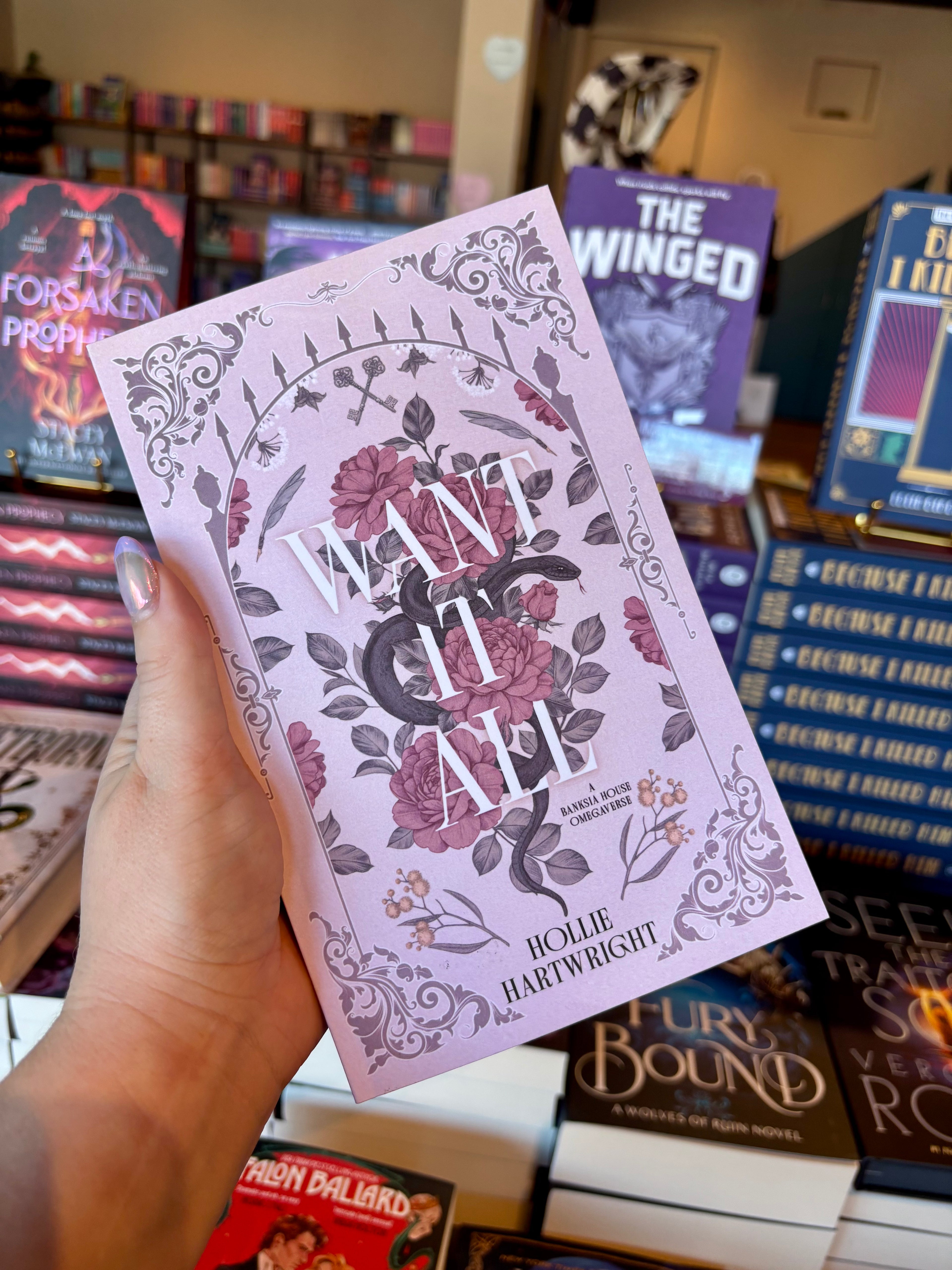 A hand holds a copy of the book 'Want It All' by Hollie Hartwright in a bookstore. The book cover is light pink with dusty pink roses, grey leaves, and black decorative flourishes, with the title 'WANT IT ALL' in large white letters.