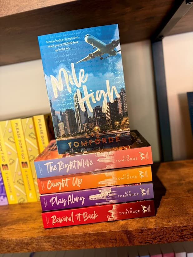 A stack of romance novels by Liz Tomporde, including 'Mile High', 'The Right Mile', 'Caught Up', 'Play Along', and 'Rewind It Back', displayed on a wooden bookshelf.