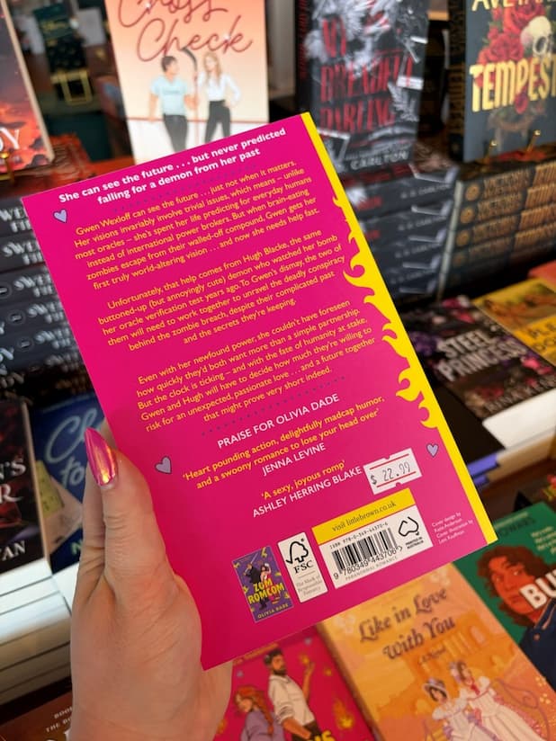 A hand with pink nail polish holds the back of a copy of 'World's Okayest Oracle (Reluctantly) Seeks Demon' by Olivia Dade, featuring a bright pink cover with a yellow spine, and yellow and white text.