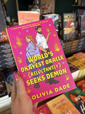A hand with pink nail polish holds a copy of 'World's Okayest Oracle (Reluctantly) Seeks Demon' by Olivia Dade, featuring a couple standing back-to-back and looking at each other over their shoulders. She has long red braided hair and is brandishing a knife, he is wearing corporate attire and has small horns poking through his hair while flames manifest from one of his hands.
