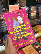 A hand with pink nail polish holds a copy of 'World's Okayest Oracle (Reluctantly) Seeks Demon' by Olivia Dade, featuring a couple standing back-to-back and looking at each other over their shoulders. She has long red braided hair and is brandishing a knife, he is wearing corporate attire and has small horns poking through his hair while flames manifest from one of his hands.
