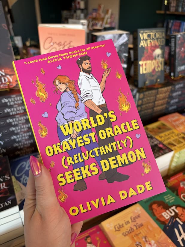 A hand with pink nail polish holds a copy of 'World's Okayest Oracle (Reluctantly) Seeks Demon' by Olivia Dade, featuring a couple standing back-to-back and looking at each other over their shoulders. She has long red braided hair and is brandishing a knife, he is wearing corporate attire and has small horns poking through his hair while flames manifest from one of his hands.