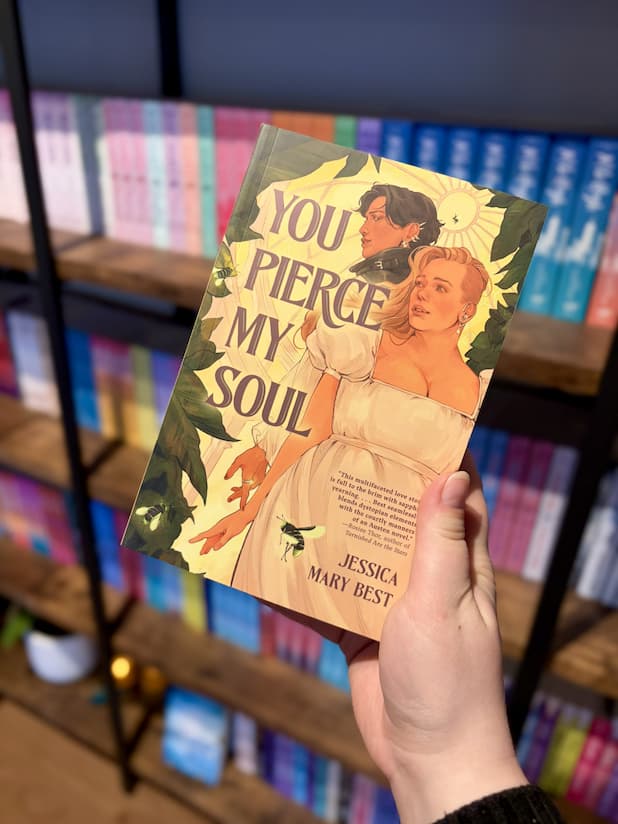 A hand holding a copy of 'You Pierce My Soul' with two women standing back-to-back with fingers barely touching, they are in a greenhouse surrounded by leaves.