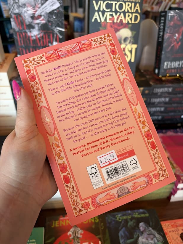 A hand with pink nail polish holds the back of a copy of 'You'll Be the Death of Me' by Holly Michelle, featuring a peach background, purple and red text, and a floral border. 