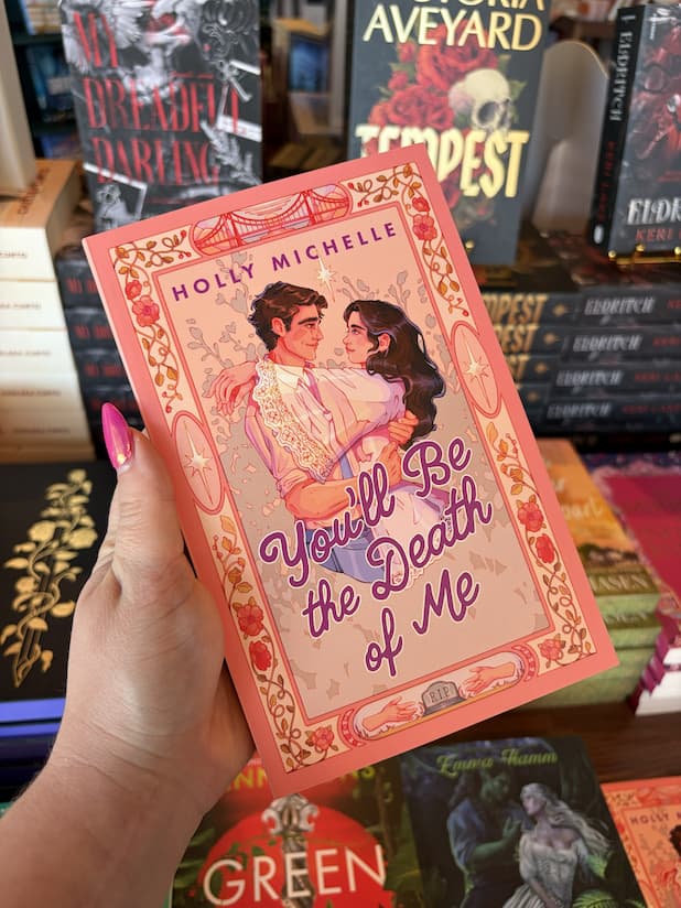 A hand with pink nail polish holding a copy of 'You'll Be the Death of Me' by Holly Michelle with a couple embracing. He's wearing corporate attire and she's translucent like a ghost. They are surrounded by a border with decorative elements.