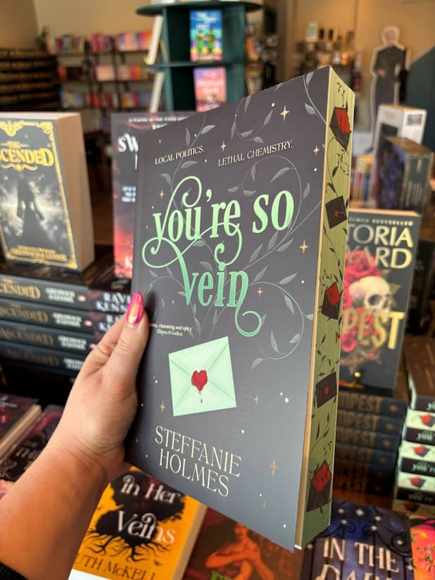 A hand with pink nail polish holds a copy of 'You're So Vein' by Steffanie Holmes, featuring a black cover with green vines and a letter with a heart-shaped wax seal, with matching sprayed edges.